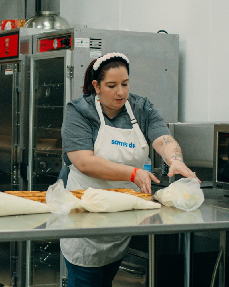 Bridgette Anderson stands at a stainless steel table in a commercial kitchen arranging large pastry bags filled with cream or frosting.