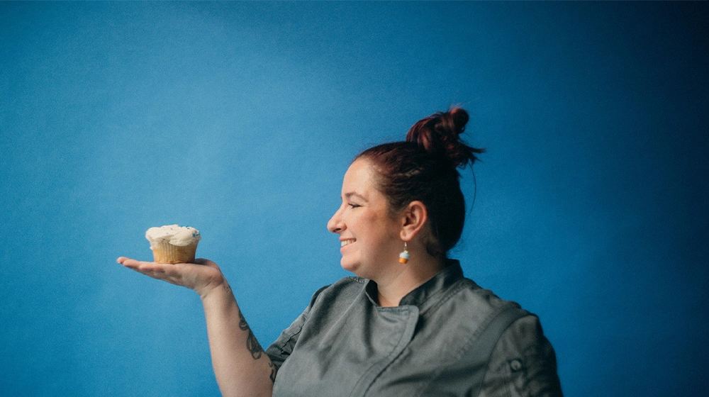 Sam's Club bakery associate Bridgette Anderson holds a cupcake in the palm of her hand, standing against a blue background.