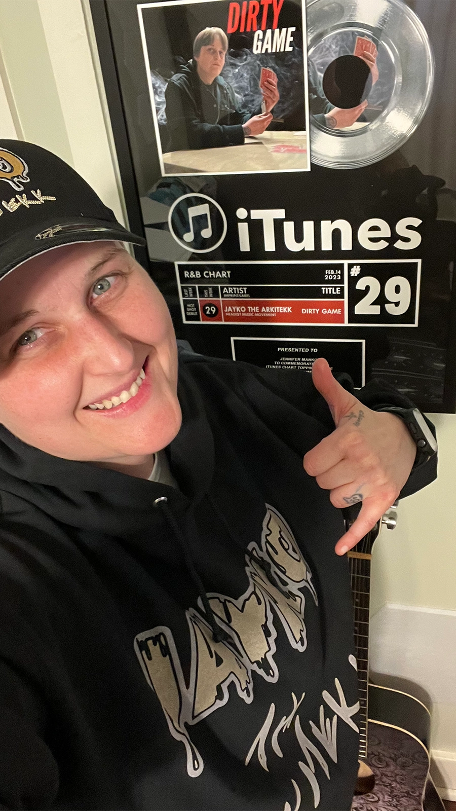 Jayko the ARkiTekk poses in front of an award from iTunes for her single, "Dirty Game," smiling at the camera.