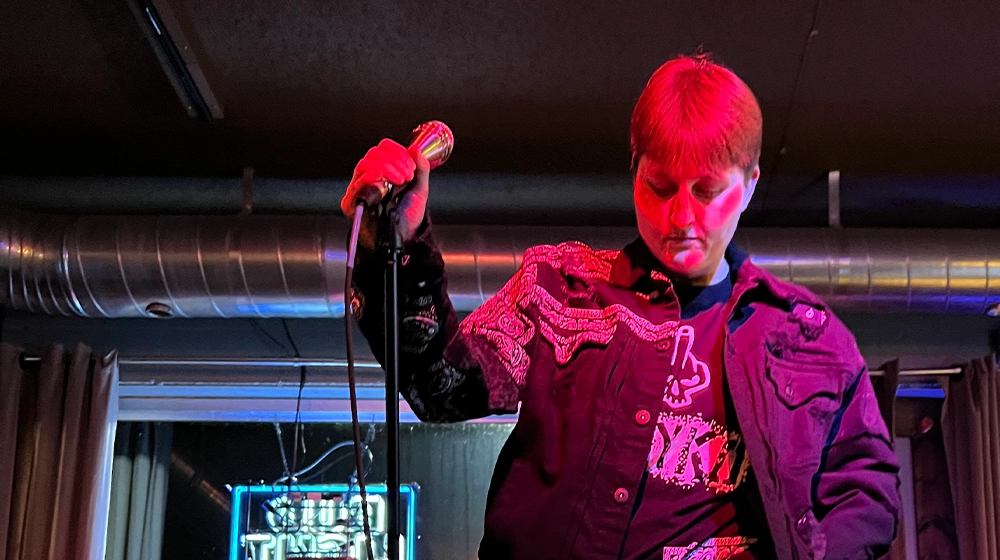 Jayko the ARkiTekk holds a microphone under neon stage lights.