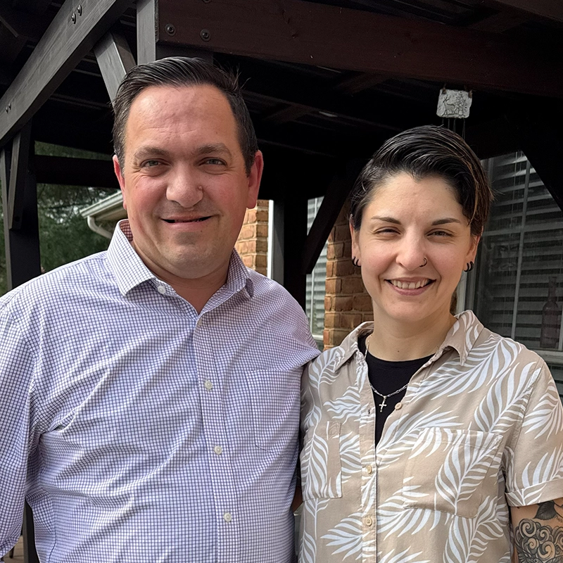 Two adults stand side by side, posing for a photo in an outdoor setting. The background features a brick wall and wooden structure, suggesting a residential or casual environment. One person wears a patterned button-up shirt with a visible cross necklace, while the other wears a checkered shirt. The overall mood is relaxed and informal.