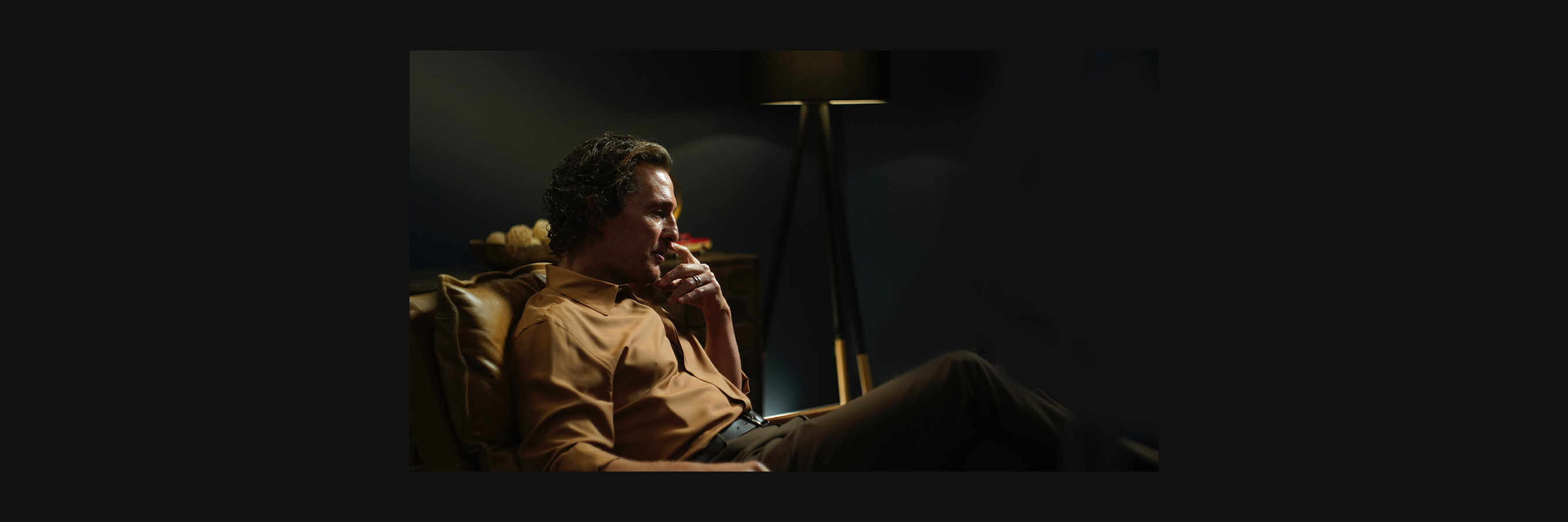 Matthew McConaughey is seated in a comfortable chair in a dimly lit room, creating a cozy and contemplative atmosphere. A modern floor lamp provides soft lighting, enhancing the warm tones of the setting.