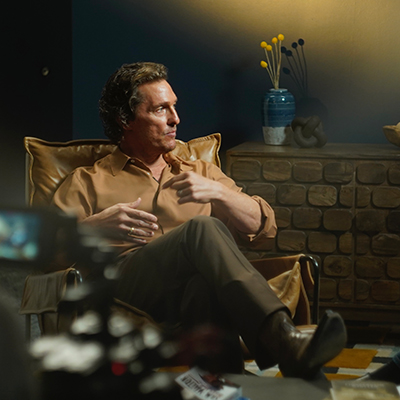 Matthew McConaughey sits in a leather armchair during an interview in a warmly lit studio. The background features a brick wall, decorative vase with flowers, and a bowl of items on a shelf. The setting includes professional recording equipment and a relaxed atmosphere.