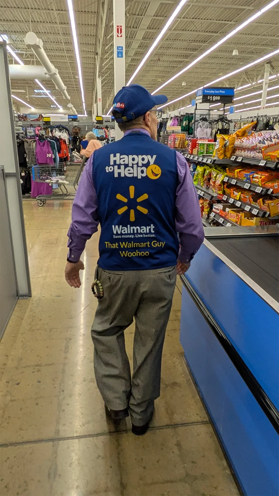Steven Harris walks down a checkout aisle with his back to the camera. He is wearing his blue Walmart vest that identifies him as "That Walmart Guy."