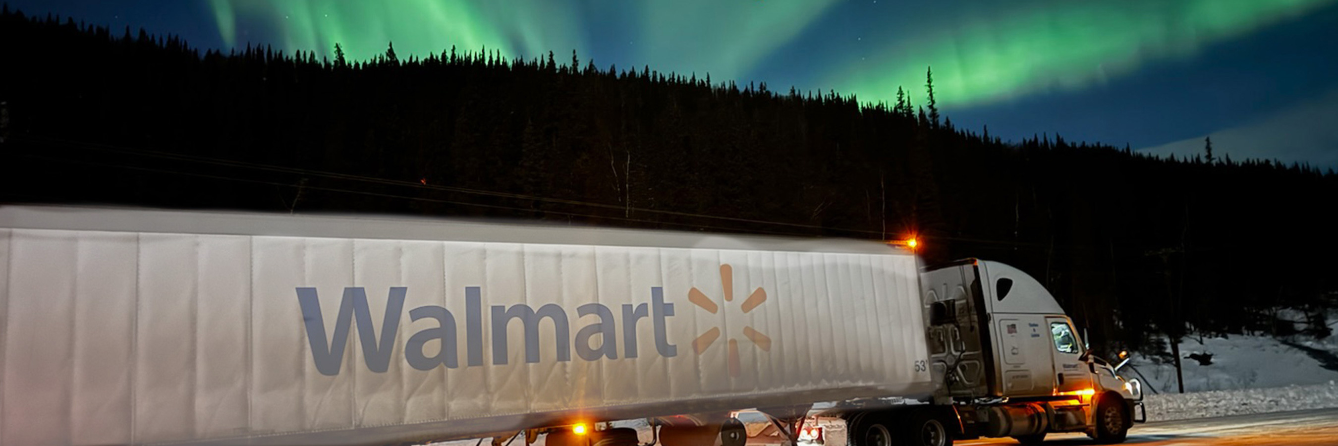 A Walmart semi truck is parked or driving along a snowy road at night, with the aurora borealis illuminating the sky above.