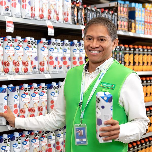 Bodega Aurrera associate German is wearing a green vest and is holding a carton in an aisle. The shelves are stocked with various cartons in organized product displays.