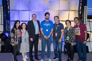 A group of individuals standing on a stage with a modular backdrop featuring white panels and blue lighting. Ankush wears a blue shirt with visible text 'Integrity' and a medal. Others are dressed in business casual and event lanyards are visible.