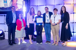 A group of individuals stands on a stage during an award ceremony. Brenda is holding a certificate, while others are dressed in formal and semi-formal attire. The setting features bright stage lighting and a geometric backdrop.
