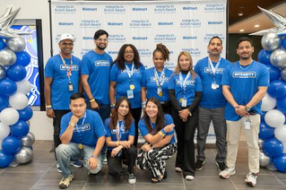 A group of individuals wearing blue 'Integrity' t-shirts pose in front of a branded Walmart backdrop. The setting includes decorative blue and white balloon arrangements and visible text such as 'Integrity in Action Award' and 'Walmart'. 