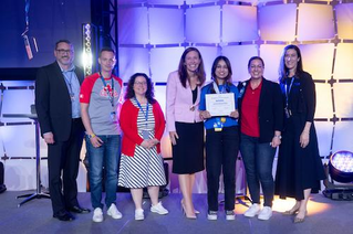 A group of individuals stands on a stage during an award ceremony. Jashandeep is holding a certificate, while others are dressed in formal and semi-formal attire. The setting features bright stage lighting and a geometric backdrop.