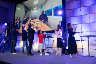 A group of people stands on a stage, clapping while a large screen displays a virtual meeting with participants. The individuals on stage include men and women of varying ages, dressed in business casual attire.