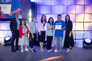 A group of individuals stands on a stage during an award ceremony. Ouyang is holding a certificate, while others are dressed in formal and semi-formal attire. The setting features bright stage lighting and a geometric backdrop.