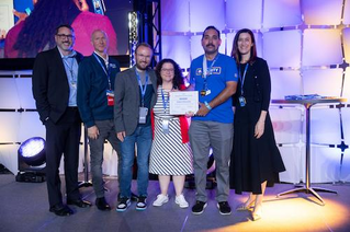 A group of individuals stands on a stage during an award ceremony. Robert is holding a certificate, while others are dressed in formal and semi-formal attire. The setting features bright stage lighting and a geometric backdrop.