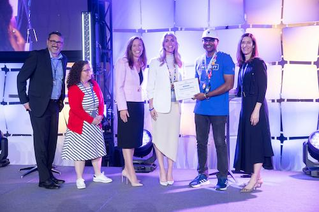 A group of individuals stands on a stage during an award ceremony. Stalin is holding a certificate, while others are dressed in formal and semi-formal attire. The setting features bright stage lighting and a geometric backdrop.
