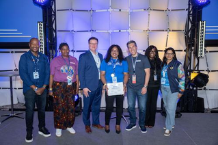 A group of individuals stands on a stage with a modern panel backdrop, illuminated by blue lighting. Thuliswa holds a certificate, while others are dressed in formal and semi-formal attire.
