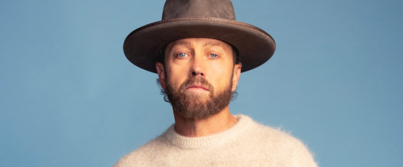 Musical artist TobyMac stares into the camera. He is wearing a wide-rimmed hat and an off-white sweater, against a sky blue background.